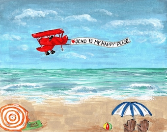 May include: A red biplane flies over a beach with a banner that reads "OCD is my happy place". The beach has a blue and white striped umbrella and a beach ball.