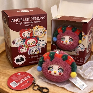 May include: A round, fuzzy, pink plush clip-on toy with a vinyl face and a multi-colored jester-style hat featuring small pom-poms. It comes in a maroon box labeled ANGELS&DEMONS vinyl face collectibles, with a matching circular red tag.