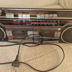 Puede incluir: Un radiocasete Sharp negro vintage con detalles plateados, que incluye un dial de radio, cubiertas de casete y altavoces. El radiocasete tiene un cable de alimentación enchufado y descansa sobre una superficie de color neutro. Dispositivo de audio retro.
