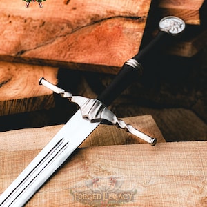 May include: A steel sword featuring a long, double-edged blade with a central fuller, a black grip, and a silver-toned crossguard with a twisted design. The pommel is circular and metallic. The text FORGED LEGACY is visible in the image.