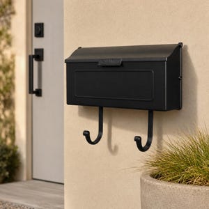 May include: A matte black wall-mounted mailbox featuring a rectangular body with a hinged lid and a small sunburst detail on the handle. Two curved hooks extend downward from the base of the mailbox, providing space for hanging items.