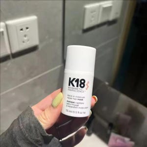 May include: A white cylindrical bottle of K18 Biomimetic Hairscience leave-in molecular repair hair mask. The label features black text and a small red molecular graphic. The bottle indicates a volume of 15 ml or 0.5 fl oz liq.