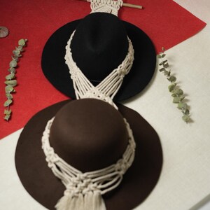 May include: Two fedora hats, one black and one brown, each with a cream macrame detail. They are displayed on a red and white surface, with a macrame hanger. Eucalyptus sprigs are placed on the side.