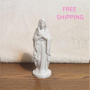 May include: A white figurine of a figure wearing a hooded robe with hands crossed over the chest. The figure stands on a circular base, featuring draped fabric details and a smooth, glossy finish. The sculpture is rendered in a solid white color.
