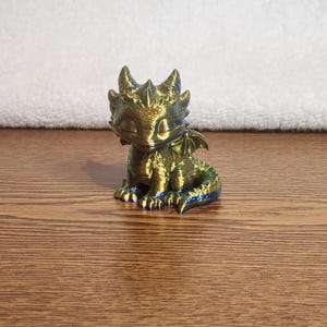 May include: A small, seated dragon figurine with a metallic, iridescent finish that shifts between shades of gold, green, and blue. The creature has closed eyes, textured scales, small wings, and a curled tail, presented in a sitting position.