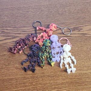 May include: A collection of five articulated skeleton keychains in various colors including metallic pink, dark purple, iridescent green, translucent light purple, and solid white, each featuring a metal ring for attachment and movable joints.