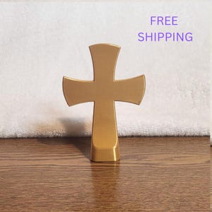 May include: A gold-colored cross with flared arms and a flat base. The cross has a smooth, solid surface and stands upright. The design features a vertical bar intersected by a horizontal bar, with all four ends widening outward in a curved shape.