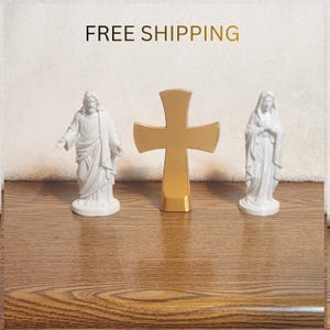 May include: Two white figurines depicting religious figures standing on either side of a central gold-colored cross. The figure on the left has arms outstretched, while the figure on the right has hands clasped at the chest. All items are smooth.