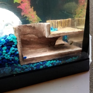May include: A tan, tiered aquarium hideout with a textured, wood-like finish sits inside a glass tank. The structure features a flat upper platform and a lower cave area, surrounded by bright blue and dark blue gravel and a pink plastic plant.