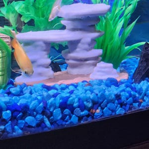 May include: An aquarium setup featuring a light purple, multi-tiered stone-like structure resting on a tan base. The tank floor is covered in bright blue gravel, and the scene includes green aquatic plants and a small orange fish swimming nearby.