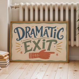May include: A rectangular framed print with a cream background featuring the text "DRAMATIC EXIT" in blue and green stylized lettering. Below the text is a terracotta-colored hand illustration pointing to the right with small star accents.