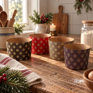May include: Four round paper baking moulds with scalloped edges, featuring various patterns including a dark brown with gold floral motifs, a red with gold floral motifs, and a tan with gold floral motifs, arranged on a wooden kitchen surface.