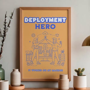 May include: A framed poster with a mustard yellow background featuring the blue text DEPLOYMENT HERO at the top and IT WORKED ON MY MACHINE at the bottom. The center shows a blue line art illustration of two people high-fiving near a computer.