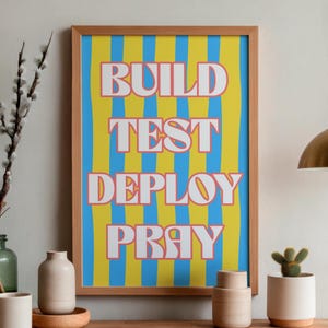 Peut inclure: Une affiche verticale encadrée présentant un fond de rayures verticales ondulées alternant entre le jaune et le bleu. Le texte "BUILD TEST DEPLOY PRAY" est imprimé en grandes lettres capitales blanches avec un fin contour rouge au centre.