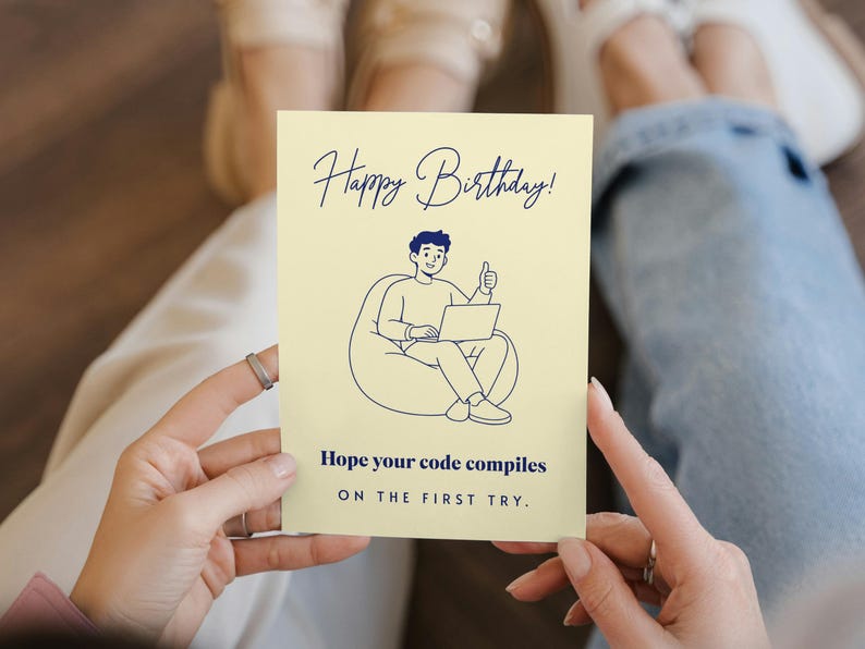 May include: A pale yellow rectangular greeting card featuring a dark blue line drawing of a person sitting in a beanbag chair using a laptop with a thumbs up. The text reads Happy Birthday! Hope your code compiles ON THE FIRST TRY.