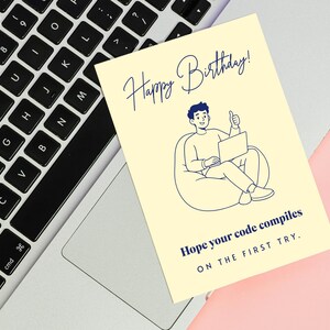 May include: A pale yellow rectangular greeting card featuring a dark blue line art illustration of a person sitting in a beanbag chair using a laptop. The text reads Happy Birthday! Hope your code compiles ON THE FIRST TRY. in dark blue ink.