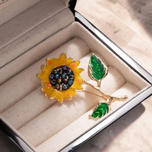 May include: A sunflower brooch with a yellow enamel flower, a dark center, and green leaves. The brooch is gold-toned and set in a black jewelry box with a cream-colored interior.