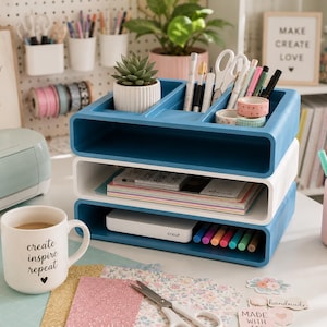 May include: A three-tiered desk organizer in blue and white holds pens, scissors, and craft supplies. A white mug with the words "create inspire repeat" sits nearby. A Cricut machine and a succulent in a white pot are also visible.