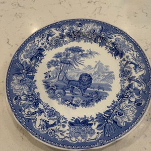 May include: A round ceramic plate featuring a blue and white transferware pattern. The center depicts a lion standing in a landscape with trees and mountains. The wide border is decorated with intricate floral motifs, including large blossoms.