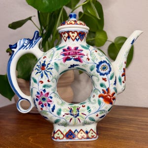 May include: A white ceramic teapot with a circular, donut-shaped body featuring a colorful floral pattern in shades of pink, blue, green, and orange. The handle is shaped like a dragon, and the lid has a matching floral design with a blue finial.