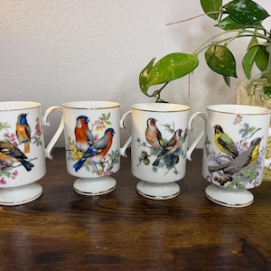 May include: A set of four white porcelain mugs with pedestal bases and gold-trimmed rims. Each mug features a detailed, colorful illustration of different birds perched on flowering branches, including finches and other small songbirds.