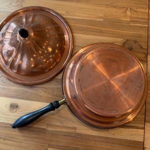 May include: A round copper pan with a long black handle and a matching circular copper lid. The lid features a fluted design and a small black knob at the center. The pan is shown upside down, revealing its smooth, polished copper base and surface.