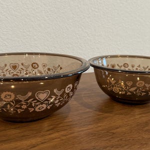 May include: Two round, translucent brown glass bowls of different sizes, each featuring a repeating white pattern of stylized birds, hearts, and floral elements encircling the exterior surface of the glass.