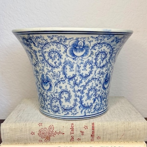 May include: A ceramic planter featuring a flared rim and a white base decorated with intricate blue floral patterns and scrolling vines. The planter sits atop two stacked books, one titled The Tales of Hoffmann and the other Rubaiyat of Omar Khayyam.