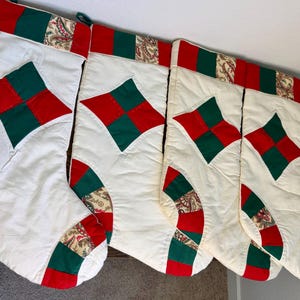 May include: Four quilted Christmas stockings in a cream fabric featuring a red and green diamond-shaped patch on the front. Each stocking has a cuff and toe section accented with a red, green, and tan paisley patterned fabric trim.