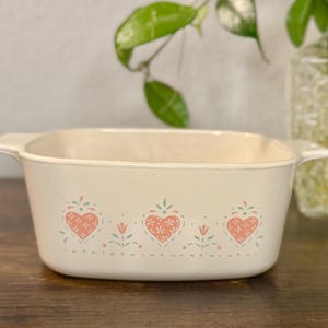 May include: A cream-colored rectangular ceramic baking dish with integrated side handles. The exterior features a repeating pattern of pink hearts and small teal floral accents arranged in a horizontal row along the center of the vessel.