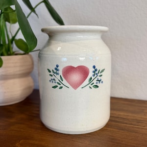 May include: A cream-colored ceramic jar with a speckled finish, featuring a central pink heart design flanked by green stems and small blue berries. The jar has a wide, rounded body and a short, slightly flared neck opening at the top.