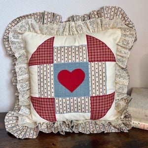 May include: A square throw pillow featuring a central blue square with a red heart appliqué, surrounded by red plaid and cream-colored floral fabric panels. The pillow is bordered by a ruffled cream lace trim and striped fabric.