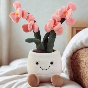 Könnte beinhalten: Ein Plüsch-Blumentopf mit einem lächelnden Gesicht, braunen Füßen und rosa Blüten mit gelben Zentren. Die Blumen haben grüne Stiele. Der Topf ist weiß.