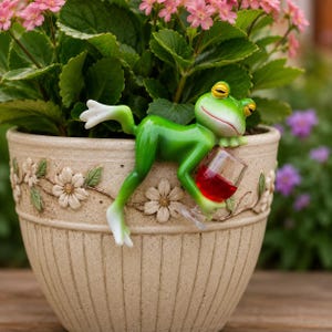Könnte beinhalten: Eine skurrile grüne Froschfigur mit gelben Augen, die ein Glas Rotwein hält, sitzt am Rand eines beigefarbenen Blumentopfs. Der Topf ist mit floralen Reliefmustern verziert und enthält rosa blühende Pflanzen. Eine lustige Gartendekoration.