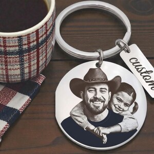 Pode incluir: Um porta-chaves circular de prata com um retrato gravado a laser a preto e branco de uma pessoa com um chapéu de cowboy, abraçada por uma criança por trás. Uma pequena etiqueta retangular de prata tem a palavra custom gravada.