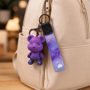 Op de afbeelding: Een paarse beren sleutelhanger met een galaxy design, met witte spikkels en X-vormige ogen. De sleutelhanger is bevestigd aan een band met een bijpassend galaxy patroon en een kleine witte wolk. De achtergrond is een beige tas.