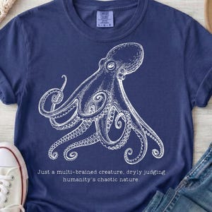 May include: A navy blue short-sleeve t-shirt featuring a white line-art illustration of an octopus. Below the graphic, the text reads: Just a multi-brained creature, dryly judging humanity's chaotic nature. The shirt has a crew neckline.