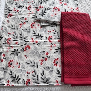 Könnte beinhalten: Ein Set Küchenwäsche mit floralem Muster und einem einfarbigen roten Handtuch. Das Blumenmuster umfasst rote Blüten, schwarze Blätter und graues Laub auf weißem Grund. Das rote Handtuch hat ein strukturiertes Gewebe.