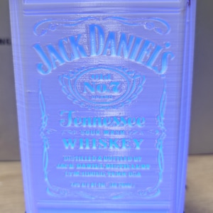 Könnte beinhalten: Eine rechteckige, durchscheinende weiße Lampe mit dem geprägten Logo und Schriftzug von Jack Daniel's Tennessee Whiskey. Die Lampe wird von innen durch ein helles blaues Licht beleuchtet und steht auf einem schwarzen Sockel mit weißem Kabel.
