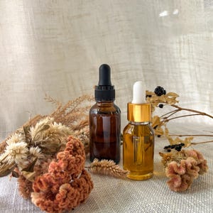 Puede incluir: Dos frascos cuentagotas de vidrio, uno de color &aacute;mbar oscuro con tapa negra y otro transparente con tapa dorada, rodeados de flores de celos&iacute;a naranja secas y hierbas beige. Los frascos son cil&iacute;ndricos y tienen pipetas de vidrio.