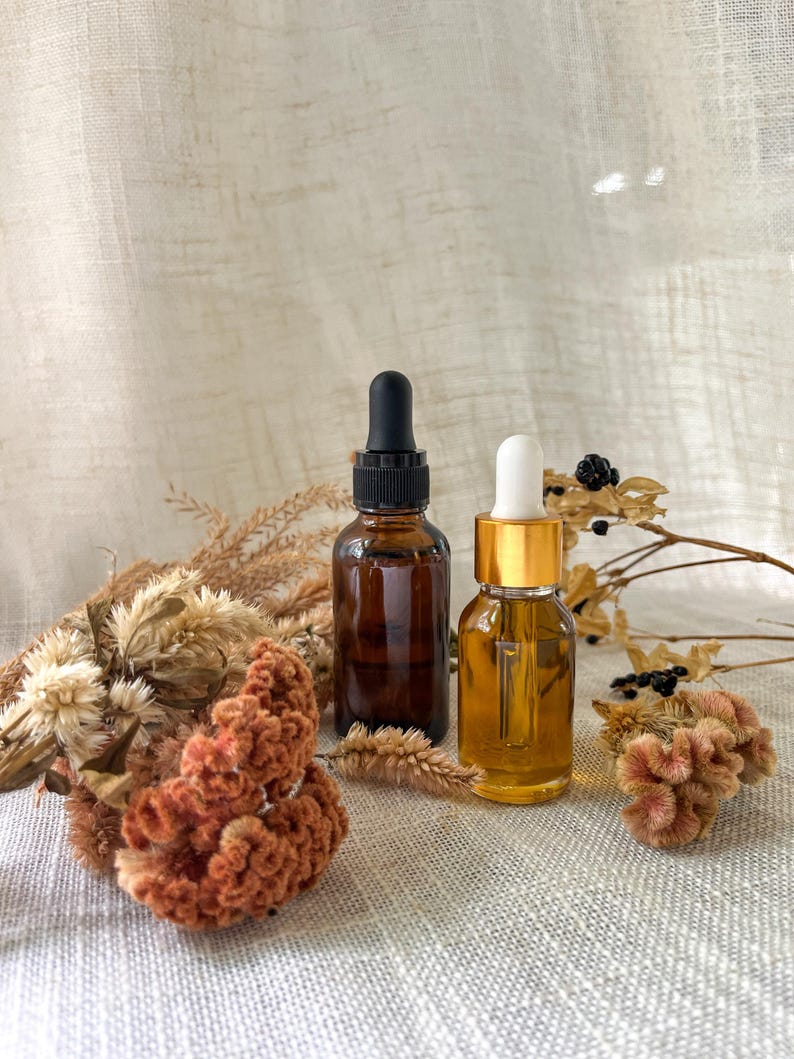 Puede incluir: Dos frascos cuentagotas de vidrio, uno de color &aacute;mbar oscuro con tapa negra y otro transparente con tapa dorada, rodeados de flores de celos&iacute;a naranja secas y hierbas beige. Los frascos son cil&iacute;ndricos y tienen pipetas de vidrio.