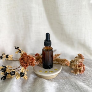 Puede incluir: Un peque&ntilde;o frasco cuentagotas de vidrio &aacute;mbar con tapa negra se asienta sobre una base de cer&aacute;mica circular. El frasco est&aacute; rodeado por tallos florales secos de color naranja y marr&oacute;n con peque&ntilde;as bayas negras, creando un arreglo natural.