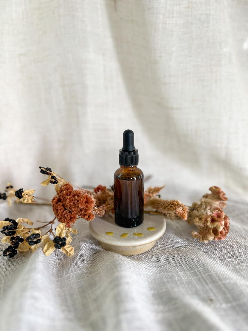 Puede incluir: Un peque&ntilde;o frasco cuentagotas de vidrio &aacute;mbar con tapa negra se asienta sobre una base de cer&aacute;mica circular. El frasco est&aacute; rodeado por tallos florales secos de color naranja y marr&oacute;n con peque&ntilde;as bayas negras, creando un arreglo natural.