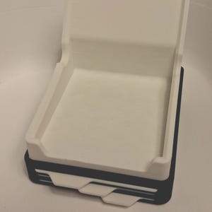 May include: A white plastic tray with a high back wall and low side walls, stacked on top of a black base. The black base features three white horizontal accent lines and a small white tab protruding from the front center of the structure.