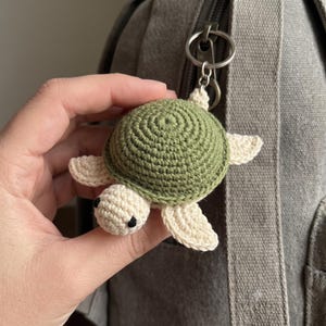 May include: A small, handmade crochet turtle keyring featuring a textured olive green shell and cream-coloured head and flippers. The turtle has two small black bead eyes and is attached to a silver-toned metal split ring for easy attachment.