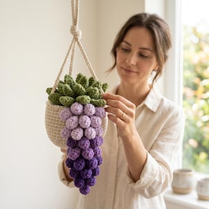 May include: A crocheted hanging planter in a cream-colored basket, featuring a green succulent top and a cascading cluster of purple and lavender crocheted spheres resembling grapes, suspended by a thick, braided cream-colored cord.