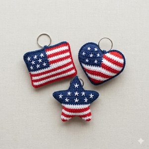 May include: Three crochet keyrings in patriotic colours. One is square, resembling a flag, another is heart-shaped, and the third is a star. Each features red and white stripes and white stars on a blue background, with a silver keyring.