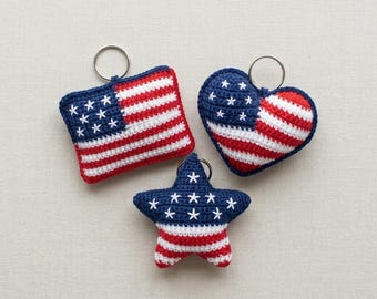 May include: Three crochet keychains in patriotic colors. One is square, resembling a flag, another is heart-shaped, and the third is a star. Each features red and white stripes and white stars on a blue background, with a silver keyring.