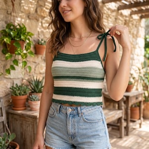 May include: A green, off-white, and light green striped crochet top with tie straps. The top has a scalloped edge and is paired with denim shorts. The image is taken outdoors with plants in the background.