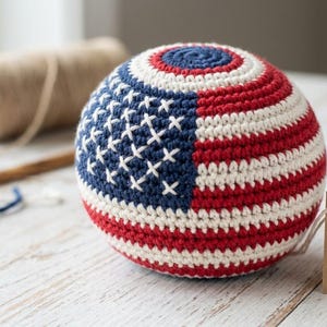 May include: A crocheted ball with a design resembling the American flag. The ball features red and white stripes, a blue field with white stars, and a tag that reads "Patriotic Ball Co."